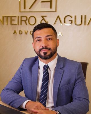 Dr. Eriverton Aguiar, de terno, sentado à mesa do escritório, com o logo da Monteiro & Aguiar ao fundo